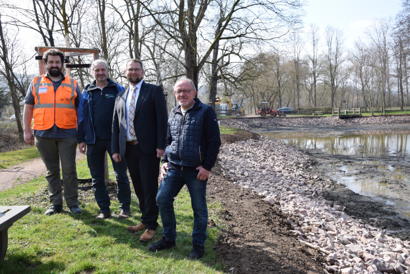 Vier Männer stehen neben einem Teich, in dem kaum Wasser mehr ist. Die Uferböschung ist mit Steinen frisch gebaut.