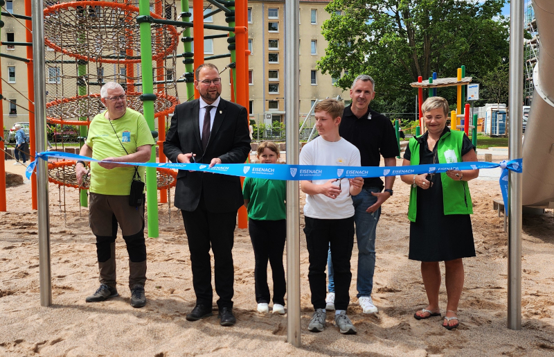 Zwei Kinder, eine Frau und drei Männer stehen auf einem Spielplatz. Sie schneiden ein blaues Band durch.